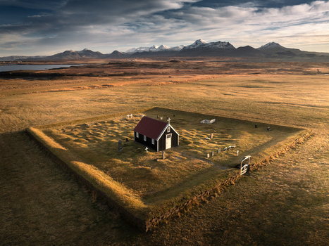 Aerial view of the historic Budakirkja black church surrounded by a stone wall in a vast grassy field with snow capped mountains in the background Iceland.
