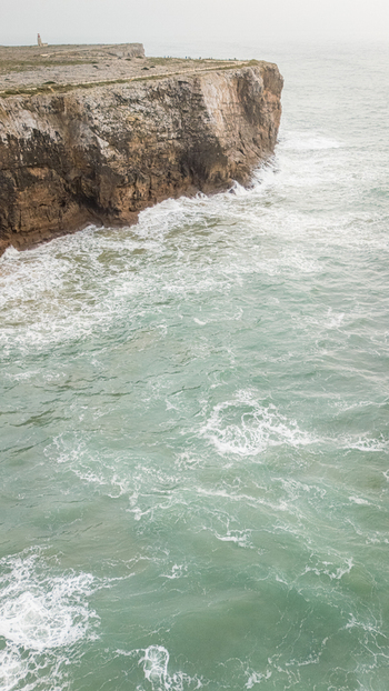 Aerial view of the rugged cliffs of Sagres Point with waves crashing against the rocks and a lighthouse in the distance Sagres Faro Portugal. Aerial view of the rugged cliffs of Sagres Point with waves crashing against the rocks and a lighthouse in the distance Sagres Faro Portugal.