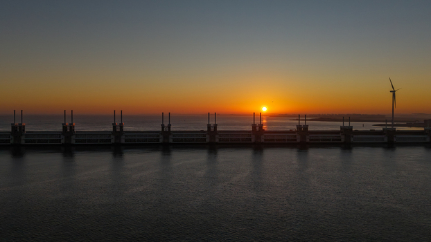 Aerial view of Deltawerken storm surge barrier with a wind turbine during a golden sunset over the North Sea.