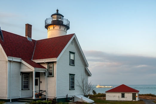 14 May 2026 View of a white lighthouse with a red roof and an attached coastal house with white siding overlooking the sea with a boat in the distance under a soft sky.