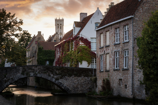 View of historic brick houses and a stone bridge over a canal with the Belfry tower in the background during sunset in Bruges Flanders Belgium.