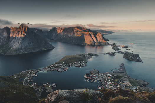 View of the dramatic Reinebringen mountain overlooking the village of Reine with red cabins deep blue fjords and rugged peaks under soft sunset light in Moskenes Nordland Norway.