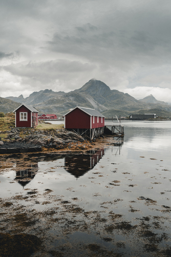 Sund Nordland Norway 04 September 2025 View of traditional red wooden rorbuer cabins on stilts along the rocky shoreline with calm water reflecting the surrounding mountains under a cloudy sky.