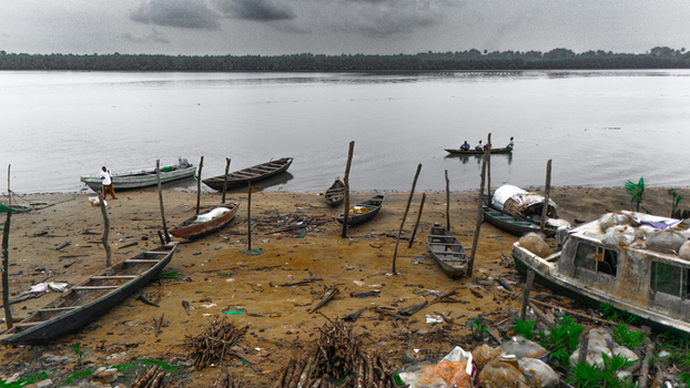 Aerial view of wooden boats moored on a muddy riverbank with people standing near the water under an overcast sky in Minima Rivers Nigeria.