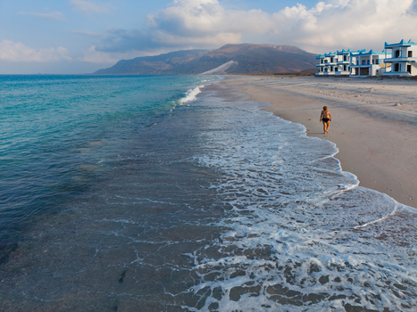 Aerial view of turquoise waves meeting the pale beach sand where a lone figure walks with blue roofed buildings standing out against the backdrop of arid mountains Ararhin Socotra Yemen. Aerial view of turquoise waves meeting the pale beach sand where a lone figure walks with blue roofed buildings standing out against the backdrop of arid mountains Ararhin Socotra Yemen.