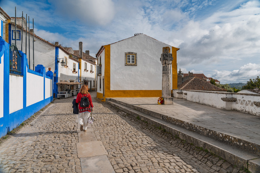 Obidos Portugal 13 May 2026 View of a woman walking along a cobblestone street in the historic town featuring traditional white buildings with yellow and blue trim and a stone pillar. Obidos Portugal 13 May 2026 View of a woman walking along a cobblestone street in the historic town featuring traditional white buildings with yellow and blue trim and a stone pillar.