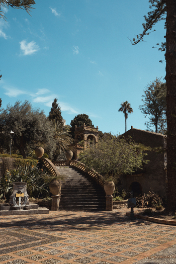 View of a stone staircase ascends through lush gardens towards an ancient structure under a sunny sky blending nature and architecture Taormina Sicily Italy. View of a stone staircase ascends through lush gardens towards an ancient structure under a sunny sky blending nature and architecture Taormina Sicily Italy.