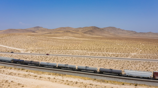 Aerial view of a long freight train with tanker cars traveling through a vast desert landscape under a clear blue sky in the United States.