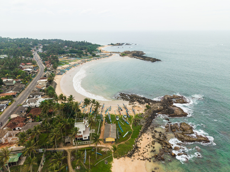 Aerial view of a curved sandy beach cove with traditional fishing boats on the shore and a coastal road Dodanduwa Southern Province Sri Lanka. Aerial view of a curved sandy beach cove with traditional fishing boats on the shore and a coastal road Dodanduwa Southern Province Sri Lanka.