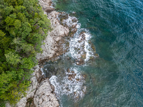 Aerial view of the rocky coastline at Razbojna City Beach with lush green trees and waves crashing against the shore in Rijeka Croatia. Aerial view of the rocky coastline at Razbojna City Beach with lush green trees and waves crashing against the shore in Rijeka Croatia.