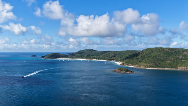Aerial view of Haslewood Island featuring lush green hills a white sandy beach and turquoise waters with a boat leaving a wake in Whitsundays Queensland Australia. Aerial view of Haslewood Island featuring lush green hills a white sandy beach and turquoise waters with a boat leaving a wake in Whitsundays Queensland Australia.