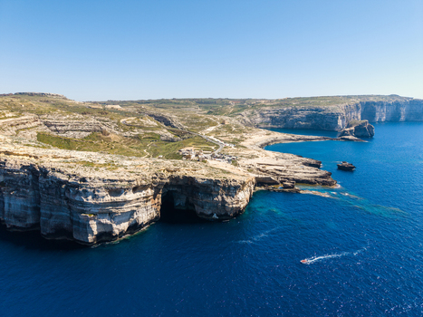 Aerial view of the rugged limestone cliffs and sea caves of Dwejra Bay with a small boat cruising on the deep blue Mediterranean water under a clear sky at Gozo Region Malta. Aerial view of the rugged limestone cliffs and sea caves of Dwejra Bay with a small boat cruising on the deep blue Mediterranean water under a clear sky at Gozo Region Malta.