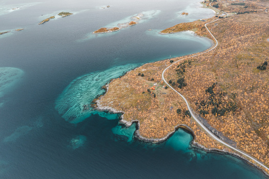 Aerial view of the rugged coastline with turquoise waters rocky shores and a winding road cutting through the autumn colored landscape in Senja Troms Norway.