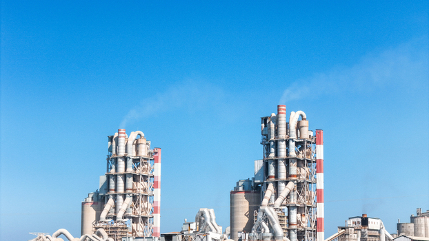 Aerial view of an industrial cement plant featuring tall silos complex piping systems and smoke stacks under a clear blue sky in Indonesia.