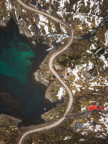 Aerial view of a winding road snaking along the rugged coastline where turquoise waters meet snow dusted rocks and quaint buildings Tind Norway.