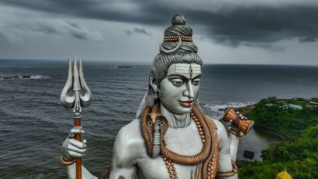 Aerial view of the massive Shiva statue at Murudeshwar Temple holding a trident and damaru set against the Arabian Sea and a dramatic cloudy sky in Murdeshwar Karnataka India.