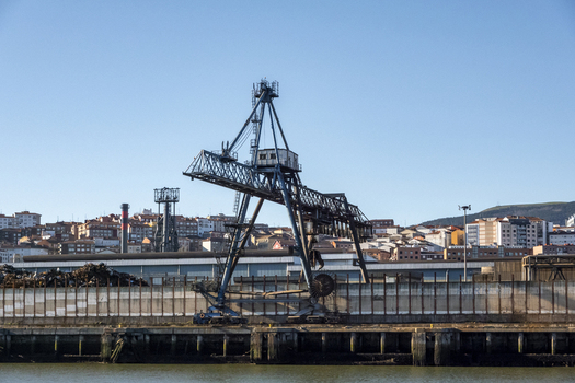 View of an industrial gantry crane standing at the waterfront with city buildings and rolling hills under a clear blue sky in Greater Bilbao Basque Country Spain. View of an industrial gantry crane standing at the waterfront with city buildings and rolling hills under a clear blue sky in Greater Bilbao Basque Country Spain.