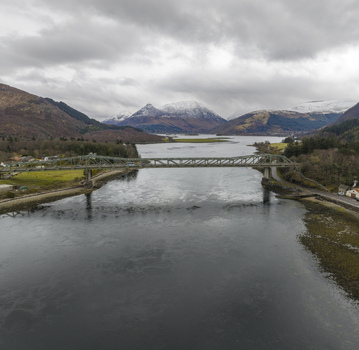Aerial view of Ballachulish Bridge spanning Loch Leven surrounded by snow capped mountains and lush green hills under a cloudy sky Ballachulish Scotland United Kingdom.