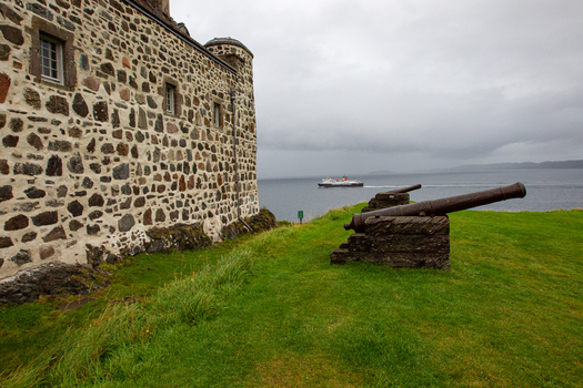 View of Duart Castle with its historic stone walls and cannons overlooking the sea where a ferry passes by under an overcast sky Isle of Mull Scotland United Kingdom.
