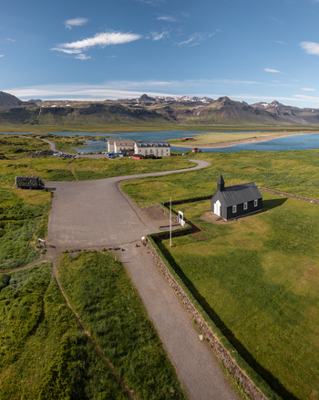 Aerial view of the iconic black Budakirkja church situated in a lush green landscape with a gravel path nearby hotel and distant mountains in Budir Snaefellsbaer Iceland. Aerial view of the iconic black Budakirkja church situated in a lush green landscape with a gravel path nearby hotel and distant mountains in Budir Snaefellsbaer Iceland.