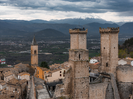 Aerial view of the medieval Caldora Castle towers and historic stone houses with snow capped mountains under a cloudy sky in Pacentro Abruzzo Italy. Aerial view of the medieval Caldora Castle towers and historic stone houses with snow capped mountains under a cloudy sky in Pacentro Abruzzo Italy.