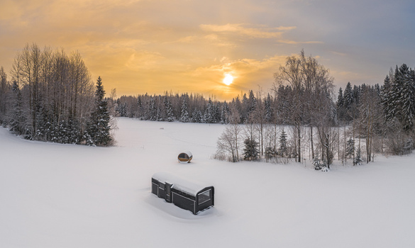 Aerial view of a small cabin and a barrel sauna in a snow covered field surrounded by a winter forest at sunset in Latvia. Aerial view of a small cabin and a barrel sauna in a snow covered field surrounded by a winter forest at sunset in Latvia.