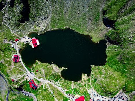 Aerial view of Balea Lake surrounded by rugged green mountain slopes winding roads and red roofed buildings in Curtea de Arges Judetul Arges Romania. Aerial view of Balea Lake surrounded by rugged green mountain slopes winding roads and red roofed buildings in Curtea de Arges Judetul Arges Romania.