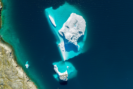 Aerial view of a large white iceberg floating in deep blue and turquoise sea water near a rugged rocky coastline in Greenland. Aerial view of a large white iceberg floating in deep blue and turquoise sea water near a rugged rocky coastline in Greenland.