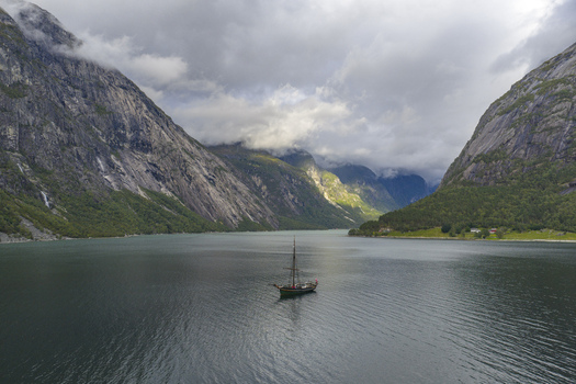 Aerial view of Hardangerfjord with a traditional sailboat navigating the calm waters surrounded by steep rocky mountains and low clouds Blurnes Vestland Norway. Aerial view of Hardangerfjord with a traditional sailboat navigating the calm waters surrounded by steep rocky mountains and low clouds Blurnes Vestland Norway.