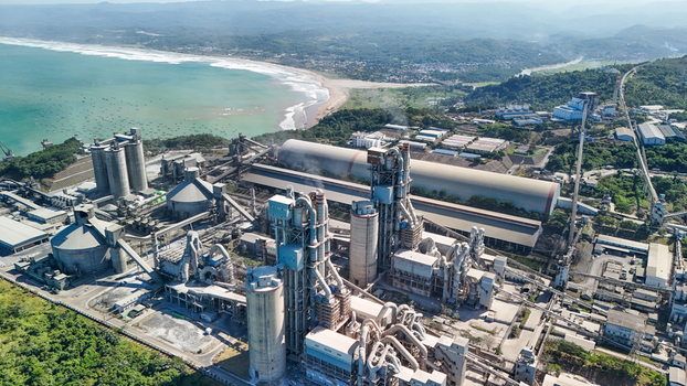 Aerial view of a large cement manufacturing plant situated along the coastline with industrial silos conveyor systems and storage buildings near lush green hills and the sea in Indonesia.