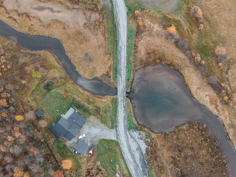 Aerial view of a house with a dark roof next to a winding stream and a road crossing a bridge in the autumn landscape in Senja Troms Norway.