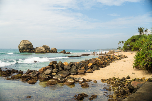 View of the sandy beach with large rock formations in the turquoise ocean and a rocky breakwater under a bright sky in Koggala Southern Province Sri Lanka. View of the sandy beach with large rock formations in the turquoise ocean and a rocky breakwater under a bright sky in Koggala Southern Province Sri Lanka.