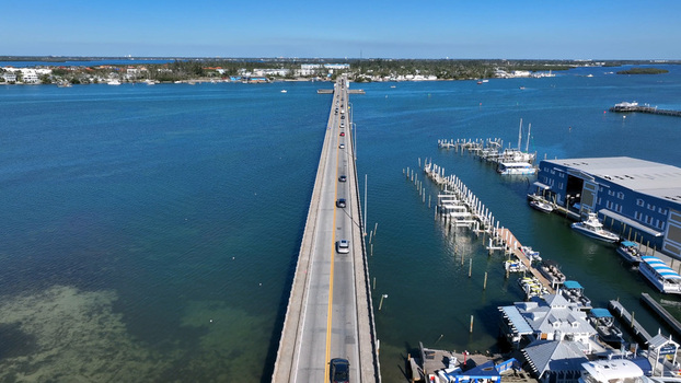 Aerial view of the Anna Maria Island Bridge spanning blue waters with cars driving across near a marina with boats and a blue building in Bradenton Florida United States.