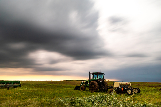 View of a green tractor working in a field under a dramatic dark stormy sky illustrating the impact of severe weather on agriculture in Oklahoma City Oklahoma United States.