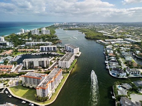 Aerial view of the Intracoastal Waterway with boats cruising past luxury waterfront apartments and lush tropical greenery under a bright sky in Boca Raton Florida United States. Aerial view of the Intracoastal Waterway with boats cruising past luxury waterfront apartments and lush tropical greenery under a bright sky in Boca Raton Florida United States.
