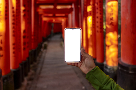 View of a hand holding a smartphone with a blank white screen in front of the red Senbon Torii Thousand Torii Gates at Fushimi Inari taisha in Kyoto Kyoto Japan.