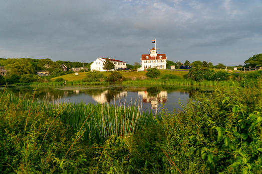View of Martha s Vineyard featuring white coastal buildings reflected in a calm pond surrounded by lush green vegetation under a cloudy sky Edgartown Massachusetts United States.