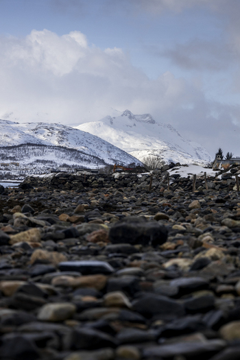 View of a rocky shoreline with snow covered mountains under a cloudy sky in Kvaloya Troms Norway. View of a rocky shoreline with snow covered mountains under a cloudy sky in Kvaloya Troms Norway.