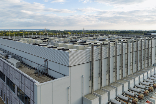 Aerial view of a large data center facility featuring numerous cooling units and ventilation pipes on the roof set against a clear sky in the United Kingdom.