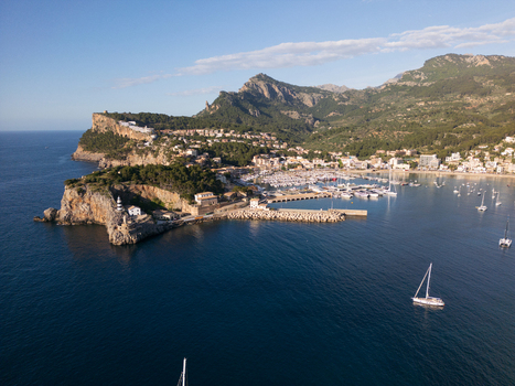 Aerial view of Port de Soller harbor with sailboats the lighthouse on the rocky cliff and the Tramuntana mountains under a clear sky Port de Soller Balearic Islands Spain.