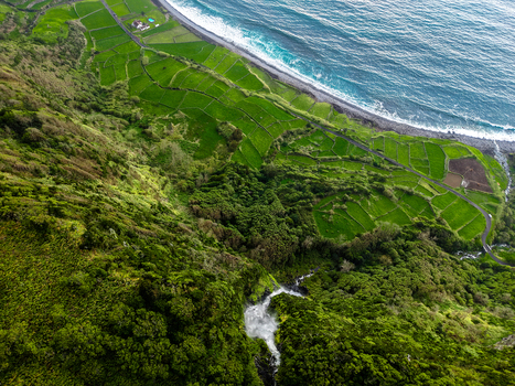 Aerial view of emerald fields meet the dark rugged coastline where crashing waves create a stark white foam against the black sand Faj&atilde; Grande Azores Portugal.