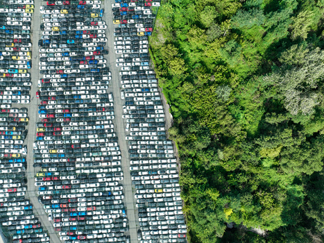 Aerial view of a large parking lot filled with rows of new cars next to a lush green forest in Turin Piedmont Italy.