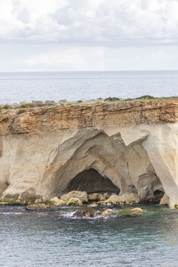 View of a large sea cave carved into a rugged limestone cliff along the Mediterranean coast under a cloudy sky Syracuse Sicily Italy.