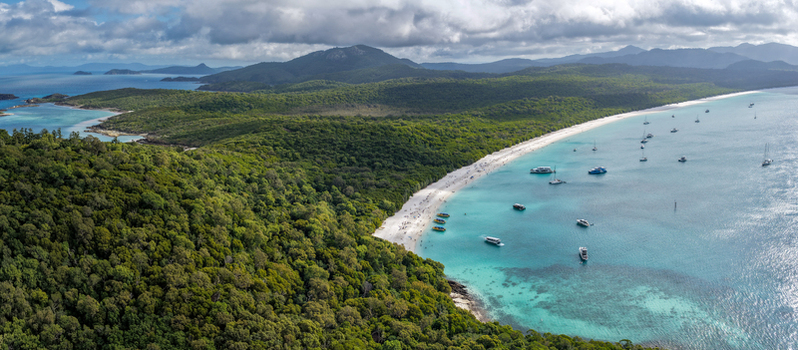Aerial view of Whitehaven Beach with its white silica sand turquoise water boats anchored in the bay and lush green forest under a cloudy sky in Whitsundays Queensland Australia.