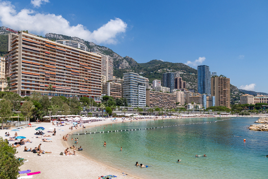 View of Larvotto Beach with people swimming and relaxing on the sand framed by modern high rise buildings and mountains under a clear blue sky in Monaco City Monaco. View of Larvotto Beach with people swimming and relaxing on the sand framed by modern high rise buildings and mountains under a clear blue sky in Monaco City Monaco.