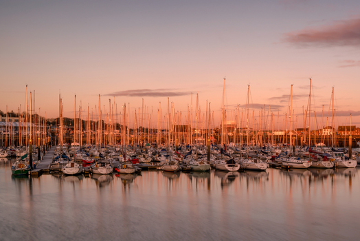 View of sailboats moored at Howth Harbour with tall masts reflecting in the calm water under a pink sunset sky Ireland. View of sailboats moored at Howth Harbour with tall masts reflecting in the calm water under a pink sunset sky Ireland.