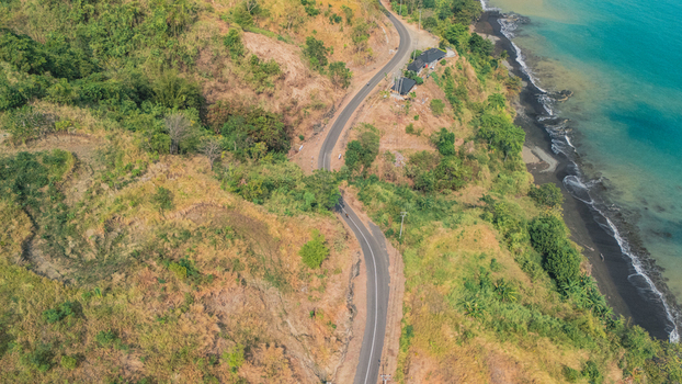 Aerial view of a winding coastal road along a cliffside with lush green vegetation and a dark sand beach bordering the turquoise ocean in Sukabumi Indonesia. Aerial view of a winding coastal road along a cliffside with lush green vegetation and a dark sand beach bordering the turquoise ocean in Sukabumi Indonesia.