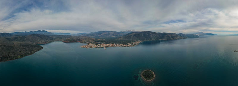 Aerial view of the coastal town of Itea featuring a small island in the foreground calm blue sea and mountainous landscape under a cloudy sky in Itea Greece.