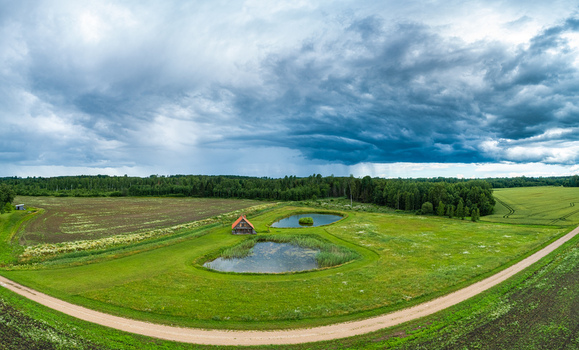 Aerial view of a small wooden house situated between two ponds in a lush green rural landscape with a dirt road and a dramatic stormy sky in Latvia. Aerial view of a small wooden house situated between two ponds in a lush green rural landscape with a dirt road and a dramatic stormy sky in Latvia.