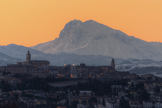 View of the historic town of Corridonia nestled against the snow capped Sibillini Mountains at sunset with warm light on stone buildings and church towers in Corridonia Marche Italy.